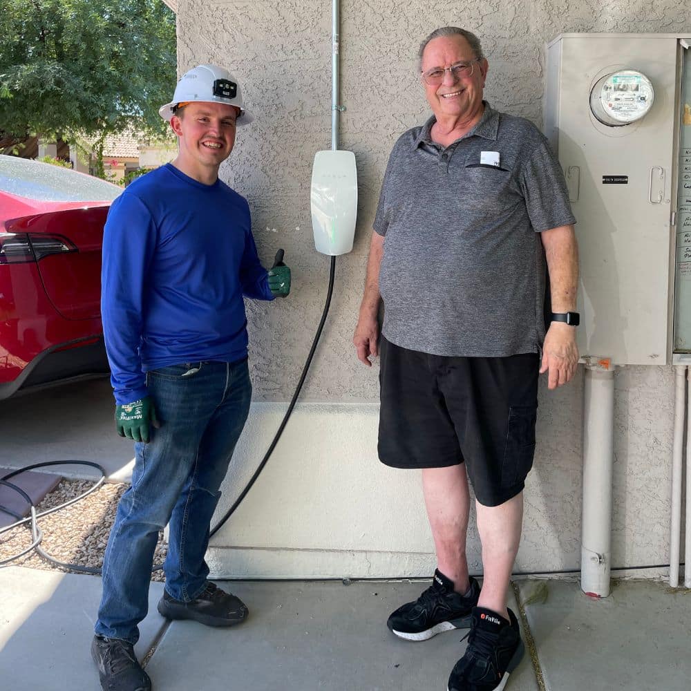 Electric vehicle charging station contractor Nick Quiros standing with a satisfied client by a Tesla home charger.